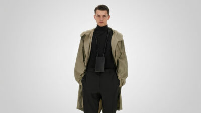 Khaki green hooded trench coat layered over a black turtleneck sweater and black tailored trousers with a leather neck pouch for THE WISHLINE luxury men's coats and trench coats collection navigation.