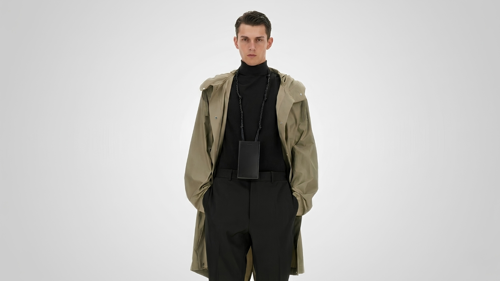 Khaki green hooded trench coat layered over a black turtleneck sweater and black tailored trousers with a leather neck pouch for THE WISHLINE luxury men's coats and trench coats collection navigation.
