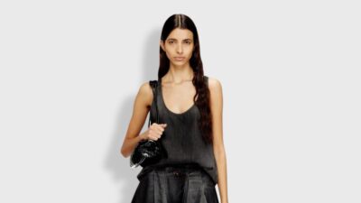 A front-facing studio shot of a model with long, dark wavy hair standing against a minimalist light grey background. She is wearing a relaxed-fit, scoop-neck tank top in a faded charcoal grey, paired with a matching dark denim bottom. A glossy, black patent leather handbag rests over her shoulder, presenting a chic and monochromatic casual luxury look.