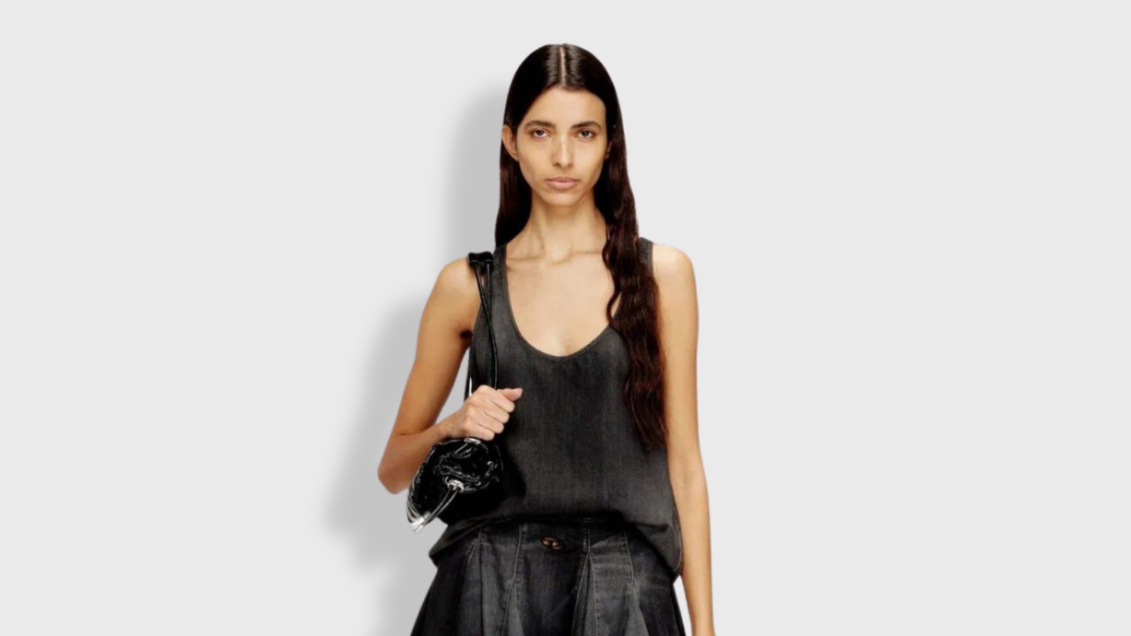 A front-facing studio shot of a model with long, dark wavy hair standing against a minimalist light grey background. She is wearing a relaxed-fit, scoop-neck tank top in a faded charcoal grey, paired with a matching dark denim bottom. A glossy, black patent leather handbag rests over her shoulder, presenting a chic and monochromatic casual luxury look.