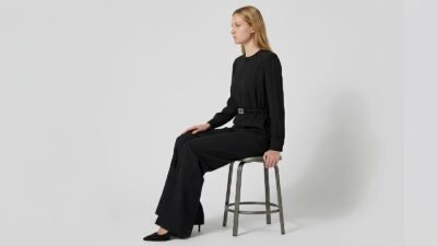 A side-profile view of a blonde model sitting on a metallic stool against a clean, minimalist white background. She is wearing a sophisticated black jumpsuit featuring long sleeves, wide-leg trousers, and a cinched waist with a subtle metallic buckle. She pairs the look with pointed black stiletto heels, embodying a sleek and modern luxury aesthetic.