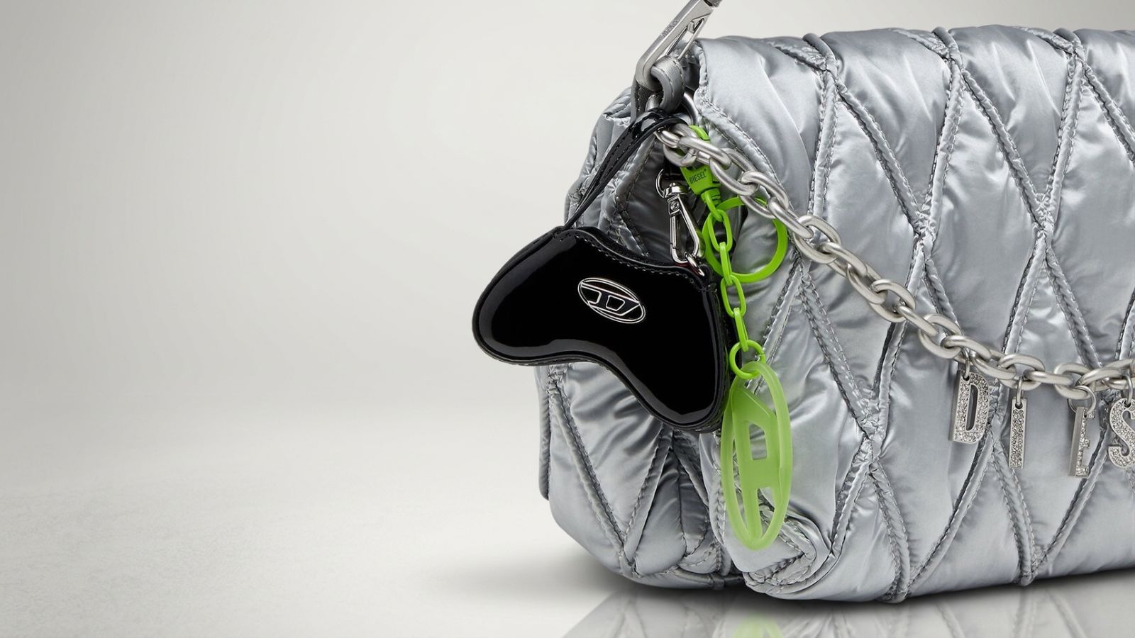 Diesel black patent leather micro bag charm and neon green 1DR logo keychain clipped to a quilted metallic silver shoulder bag for THE WISHLINE luxury keychains collection navigation.