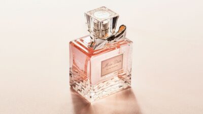 A square glass bottle of Miss Dior Eau de Toilette perfume set against a soft, shimmering beige background. The elegant bottle features a clear, cube-shaped cap, a metallic silver bow tied around the neck, and a classic rectangular label on the front, casting a gentle shadow and reflecting a warm, luxurious glow.