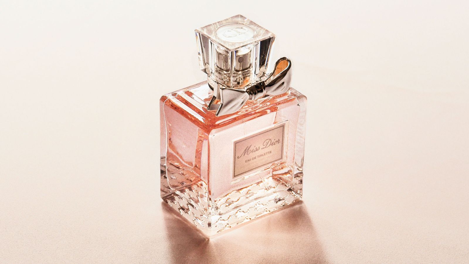 A square glass bottle of Miss Dior Eau de Toilette perfume set against a soft, shimmering beige background. The elegant bottle features a clear, cube-shaped cap, a metallic silver bow tied around the neck, and a classic rectangular label on the front, casting a gentle shadow and reflecting a warm, luxurious glow.