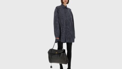 A three-quarter view of a model standing against a clean white background, showcasing an oversized, long-sleeved button-down shirt. The shirt features a dark navy or black fabric with crisp vertical white pinstripes, a pointed collar, and a relaxed, tunic-length silhouette. The outfit is paired with solid black leggings and a large, structured black leather shoulder bag