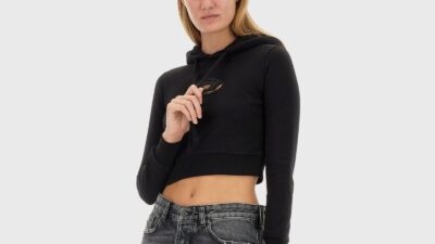 A close-up shot of a model wearing a cropped black hoodie sweatshirt against a neutral grey background. The long-sleeved sweatshirt features an oval cutout on the chest displaying a stylized "D" logo emblem, ribbed cuffs and waistband, and adjustable drawstrings held by the model's hand. She pairs the athletic streetwear look with low-rise, distressed grey denim jeans.