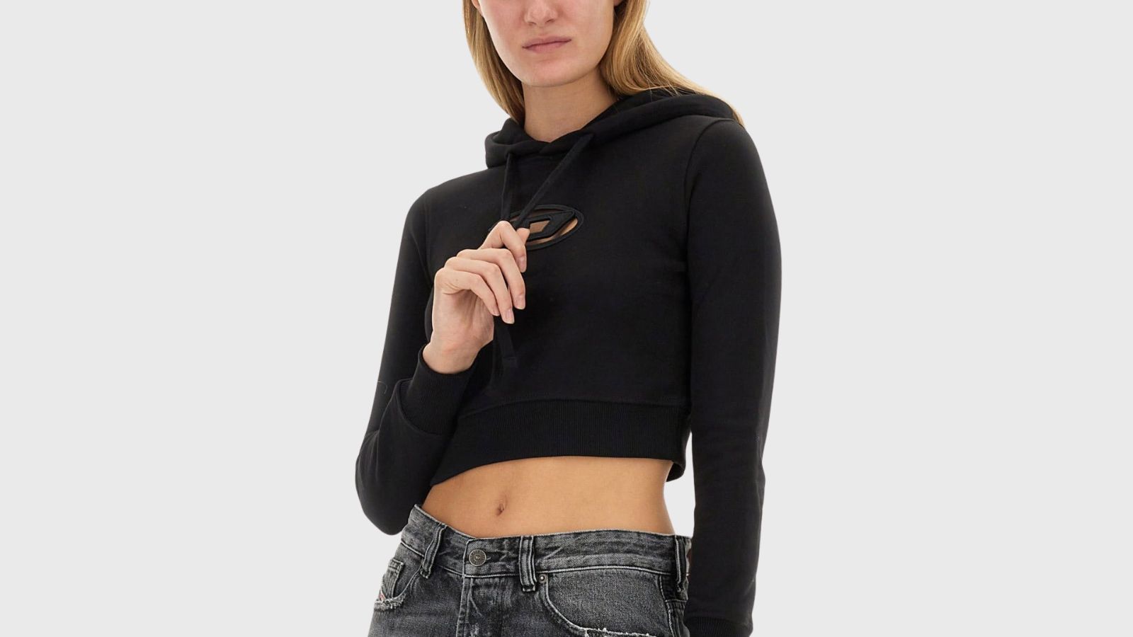A close-up shot of a model wearing a cropped black hoodie sweatshirt against a neutral grey background. The long-sleeved sweatshirt features an oval cutout on the chest displaying a stylized "D" logo emblem, ribbed cuffs and waistband, and adjustable drawstrings held by the model's hand. She pairs the athletic streetwear look with low-rise, distressed grey denim jeans.