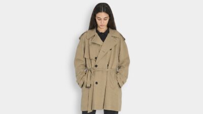 Model wearing a classic double-breasted tan trench coat with a waist belt and storm flaps for THE WISHLINE luxury women's trench coats collection navigation.