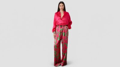 A full-length studio portrait of a model with long dark hair standing against a plain, light grey background. She is wearing high-waisted, wide-leg satin trousers featuring a vibrant, large-scale floral pattern in hot pink and green.