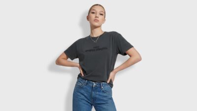A studio portrait of a model with her hair sleeked back, posing with hands on her hips against a bright white background with clean casting shadows. She is wearing a casual, washed dark grey t-shirt featuring an upside-down embossed MM6 Maison Margiela logo on the chest. The look is completed with high-waisted, classic blue denim jeans and a delicate silver necklace, showcasing a premium, effortless streetwear style.