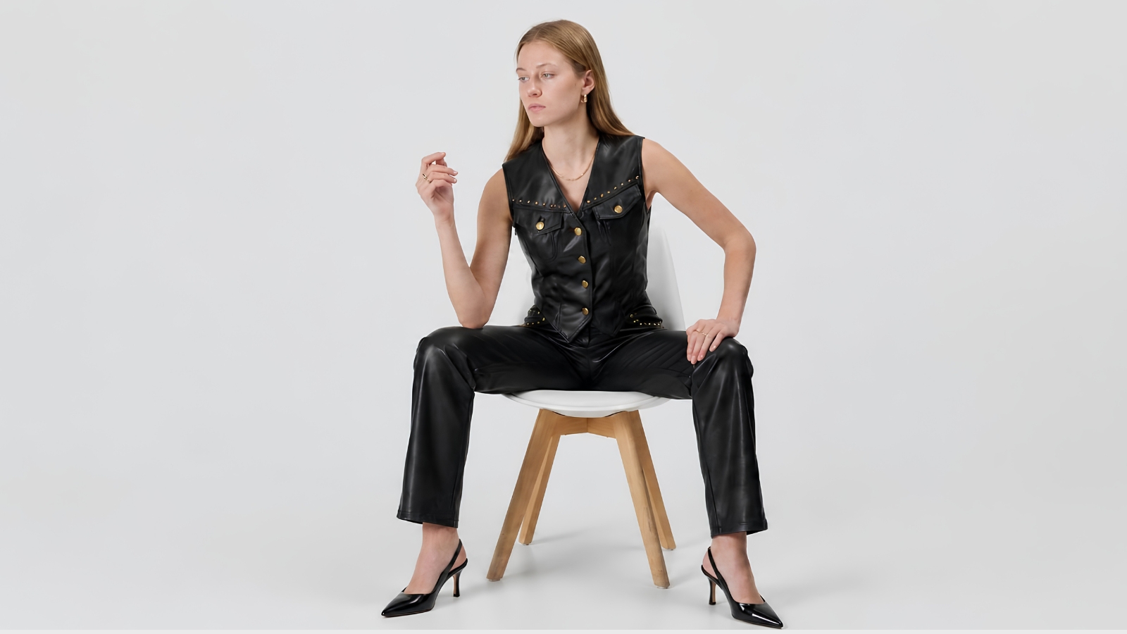 A full-length shot of a blonde model sitting forward on a white chair with wooden legs, set against a plain light grey studio background. She is wearing an edgy, sophisticated black faux leather vest featuring a V-neckline, gold-tone button closures down the front, and studded hardware details along the chest seams. The vest is styled as part of a matching set with straight-leg black leather trousers and pointed black slingback kitten heels, creating a bold, contemporary luxury look.