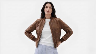 Model styling a classic brown leather bomber jacket over a white top and striped trousers for THE WISHLINE luxury women's clothing collection navigation.