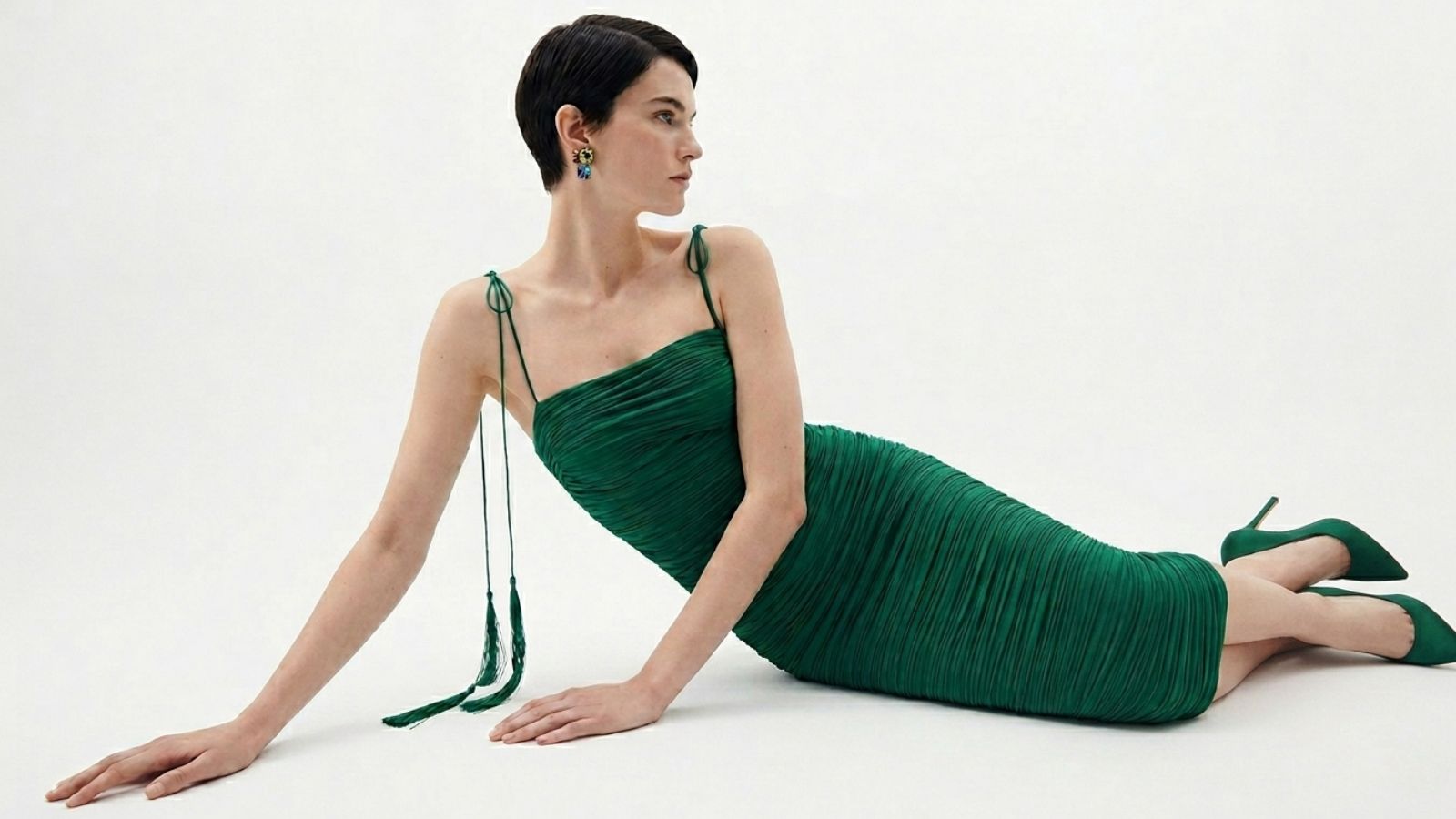 Model lounging in a sophisticated emerald green pleated midi dress with matching stiletto heels for THE WISHLINE luxury dresses navigation.