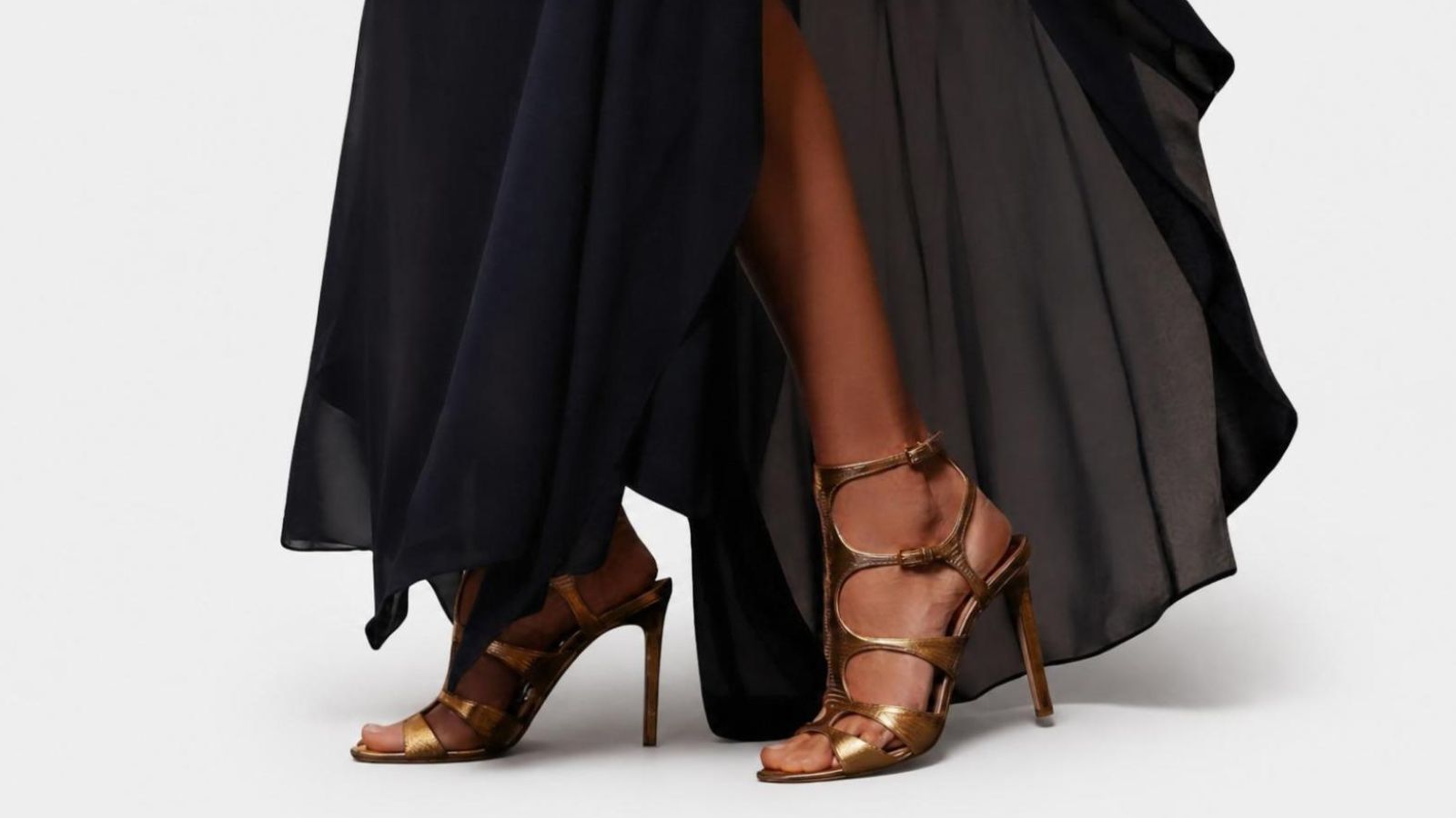 Close-up of metallic bronze strappy high-heel sandals styled with a flowing black evening skirt for THE WISHLINE designer shoes navigation.
