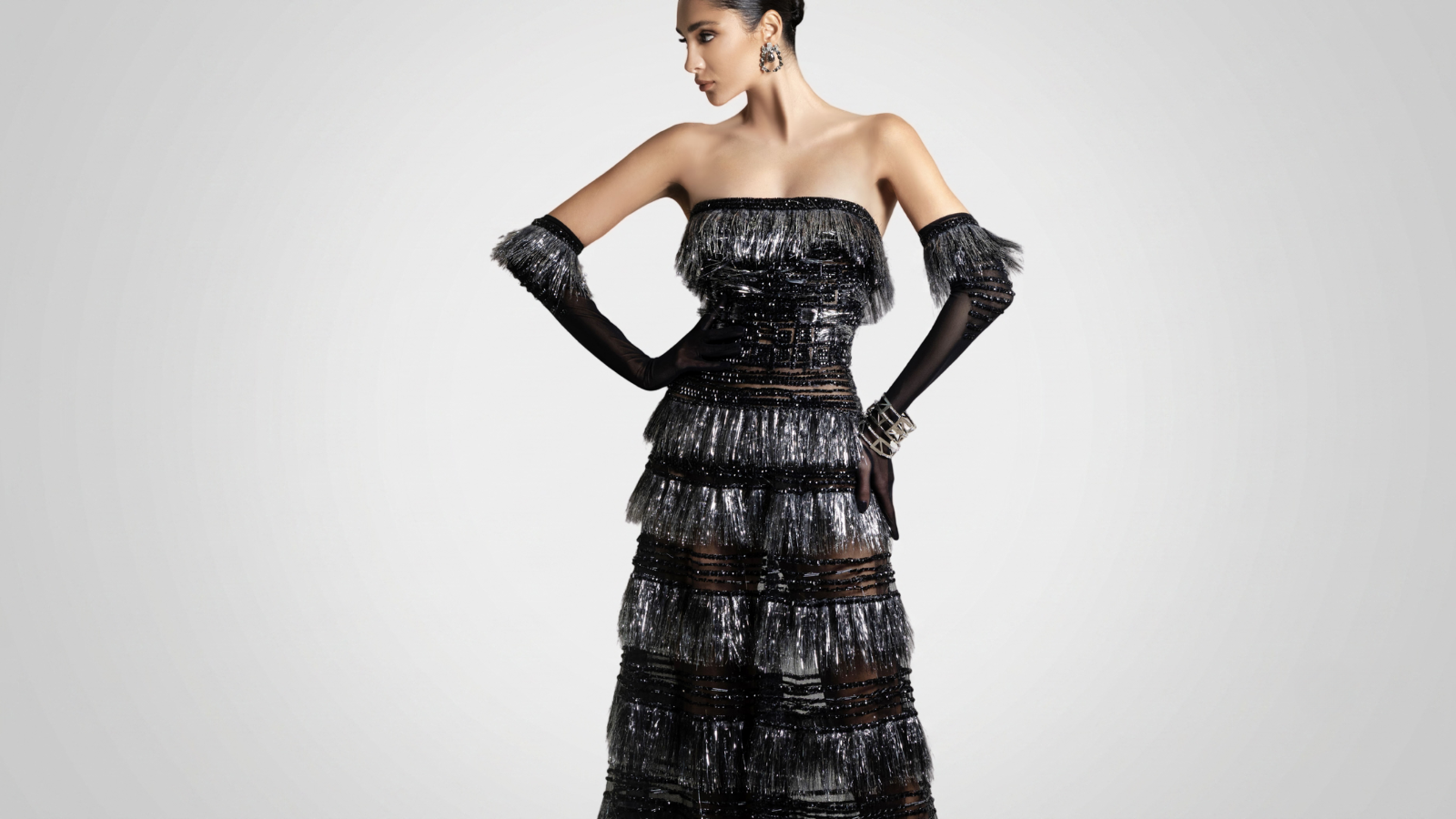 Elegant woman model in a couture strapless tiered metallic fringed black and silver gown with matching fringed opera gloves, posing against a soft gradient light grey studio background with subtle shadows.