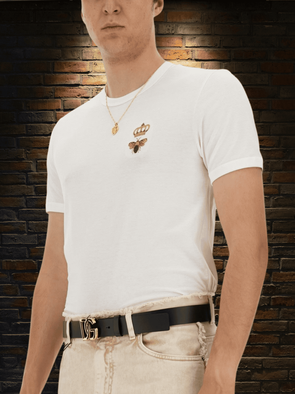 Close-up of a man wearing a white Dolce & Gabbana t-shirt with gold bee embroidery and a black DG logo belt, set against a dark brick wall background — part of the men’s collection at THE WISHLINE.