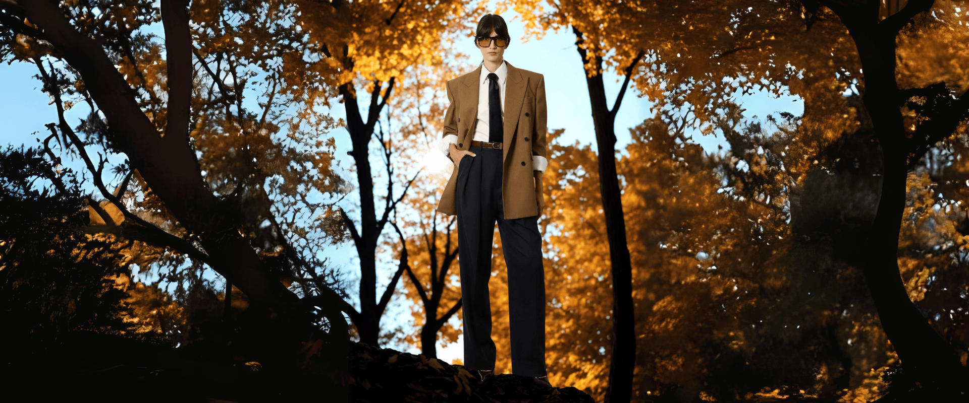 FW25 Women’s Collection – female model in a camel blazer, white shirt, black tie, and tailored trousers, standing in autumn forest setting – Fall Winter 2025 designer fashion at THE WISHLINE.