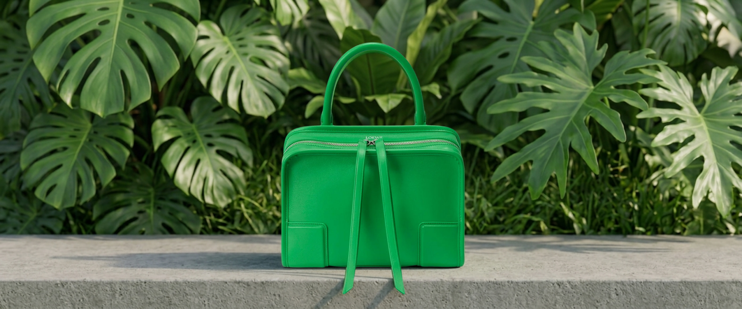 Front-facing view of the Loewe Amazona top-handle bag in vivid Kelly Green leather, resting on a textured gray concrete plinth with a dense background of lush, architectural tropical green foliage.