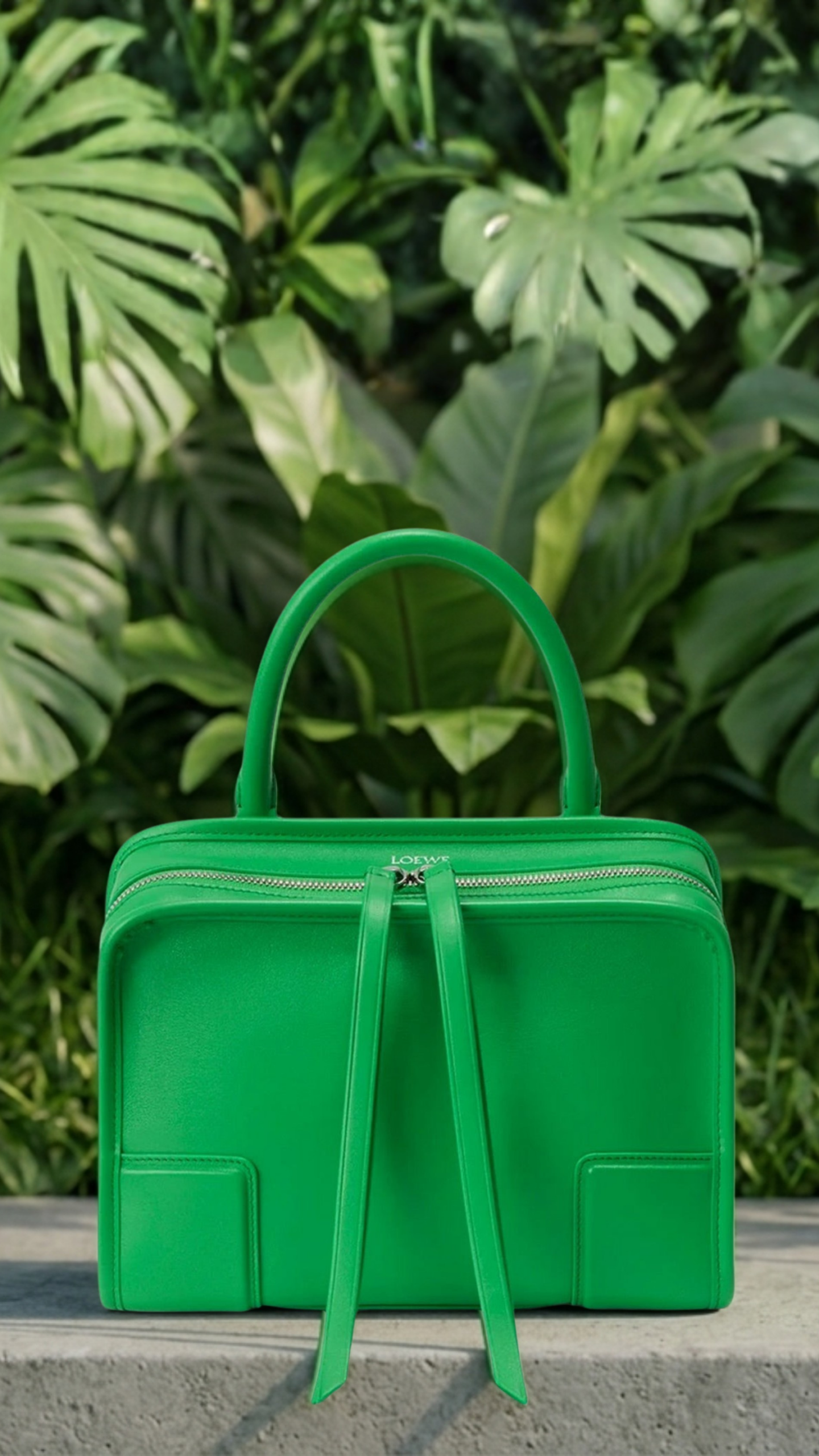 Front-facing view of the Loewe Amazona top-handle bag in vivid Kelly Green leather, resting on a textured gray concrete plinth with a dense background of lush, architectural tropical green foliage.