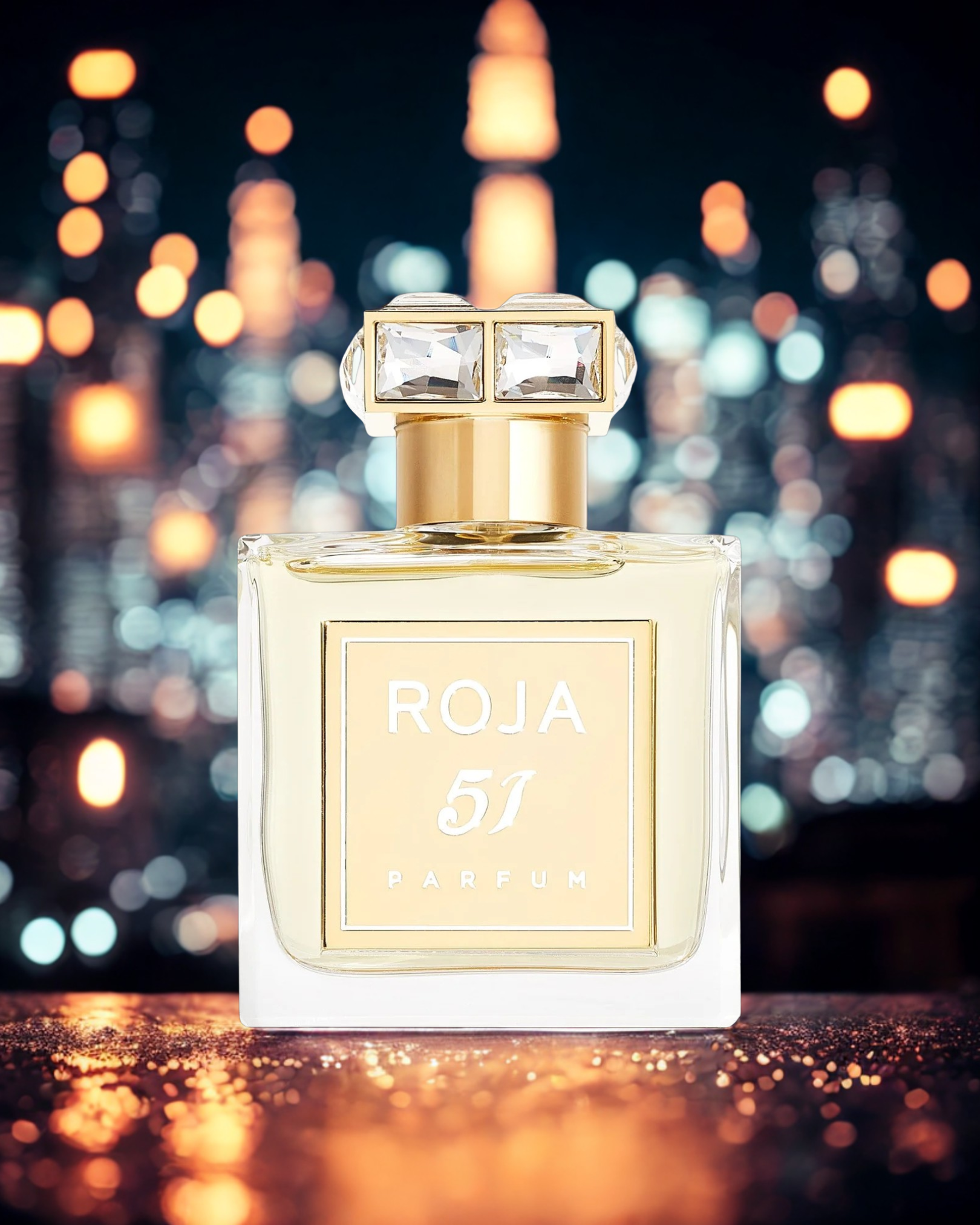 Roja 51 perfume bottle with silver crown cap placed on a reflective surface, illuminated by warm bokeh lights — representing THE WISHLINE Men’s Luxury Line Up.
