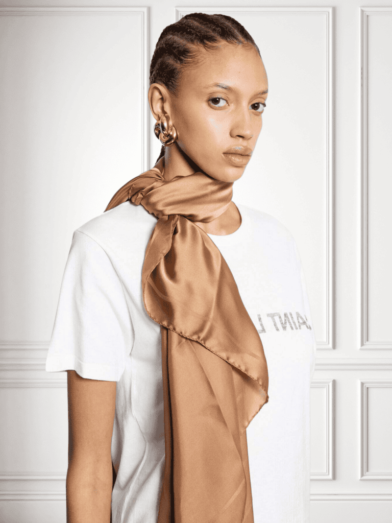Woman wearing a Saint Laurent gold silk scarf elegantly draped over a white tee, paired with statement gold earrings—luxury meets effortless style.